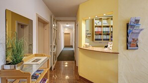 A hotel reception area with a counter, a mirror, a plant, and a hallway leading to another room.
