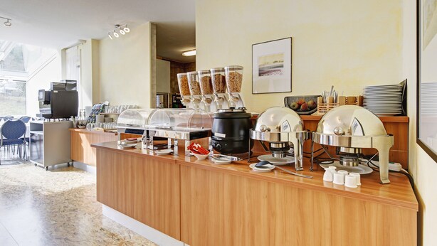 A breakfast bar with coffee machines, a buffet station with covered food containers, and a seating area with chairs.