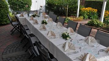 A long table set for a formal outdoor dining event, surrounded by greenery and brick flooring.
