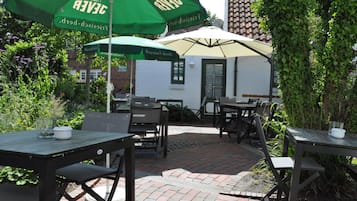 Un patio al aire libre con mesas y sillas bajo sombrillas verdes, rodeado de vegetación y un edificio de fondo.