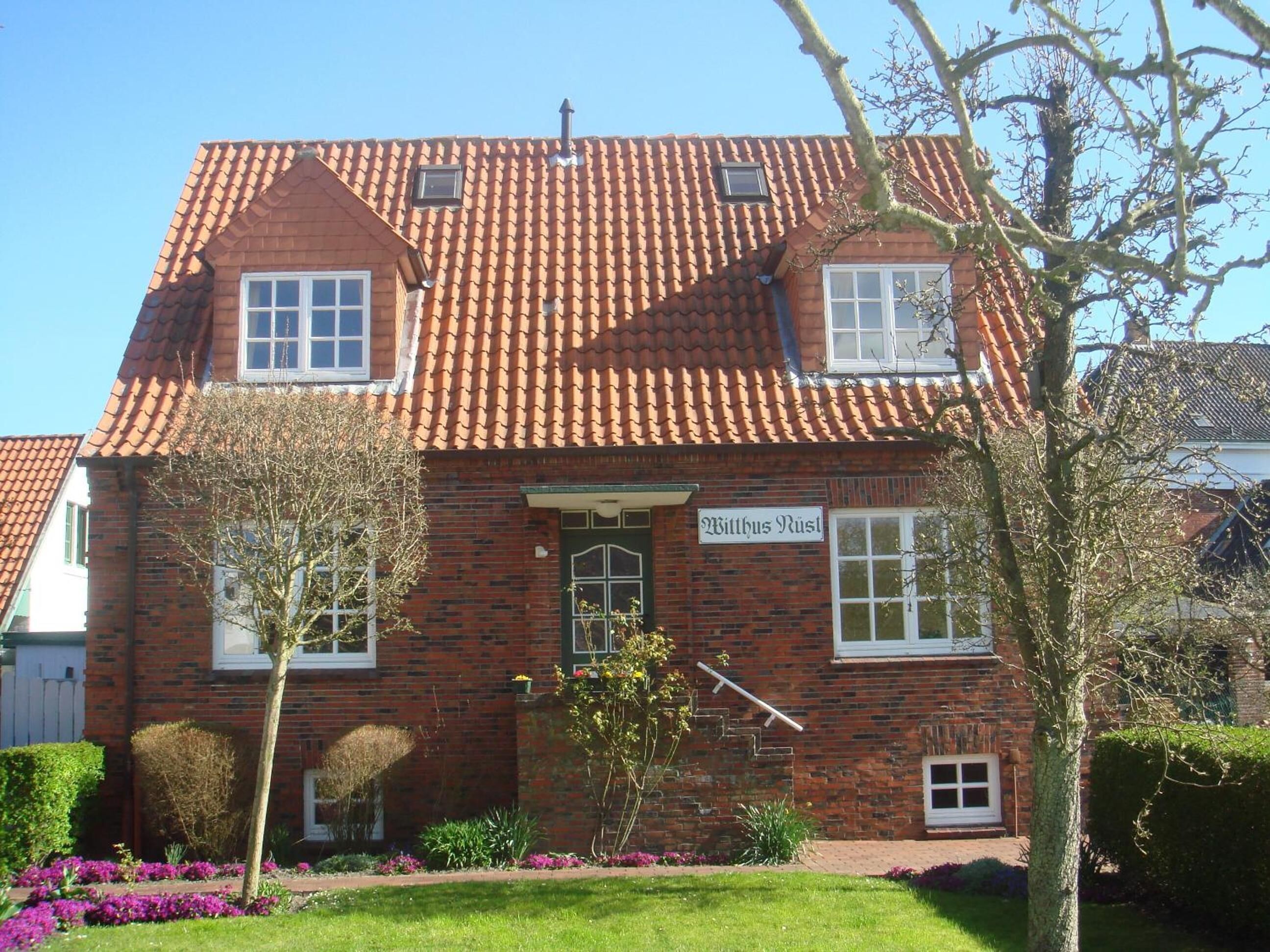 A two-story brick building with a tiled roof, a sign reading "Wittus Rust," and a well-maintained garden with flowering plants.