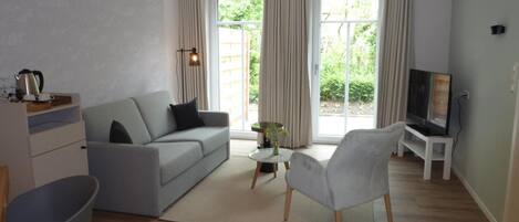 Un salón moderno con un sofá gris, un sillón blanco, un televisor sobre un mueble bajo y un ventanal grande con cortinas.