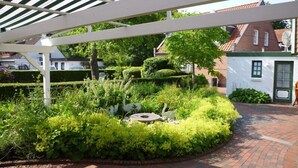 A garden with a white pergola, a brick pathway, and a small white building with a green door. - Hotel Restaurant Witthus (Krummhoern)