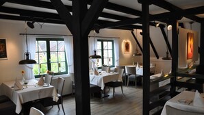A dining area with tables set for two, wooden beams on the ceiling, and large windows with curtains. - Hotel Restaurant Witthus (Krummhoern)