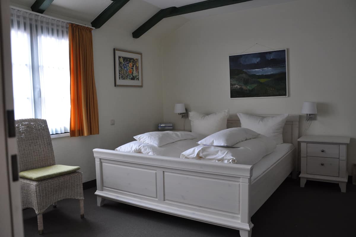 Ein Schlafzimmer mit einem weißen Bett, zwei Nachttischen, einem Sessel, einem Fenster mit Vorhängen und zwei Wandleuchten.