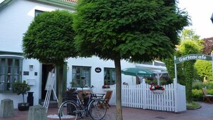 A white building with a sign reading "Gartencafe." There are two bicycles parked outside, a white picket fence, and a green umbrella. - Hotel Restaurant Witthus (Krummhoern)