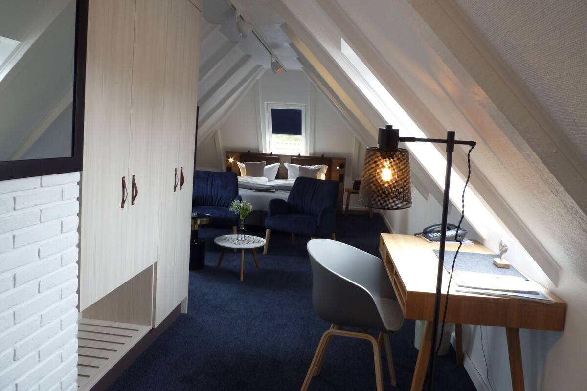 Ein gemütliches Schlafzimmer im Dachgeschoss mit schrägen Wänden, einem Bett mit Kissen, einem Schreibtisch mit Lampe und einem Stuhl.