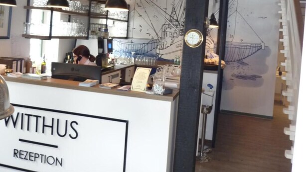 A reception area with a counter labeled "WITHUS RECEPTION," a bar area with shelves of bottles and glasses, and a large abstract wall art piece. - Hotel Restaurant Witthus (Krummhoern)