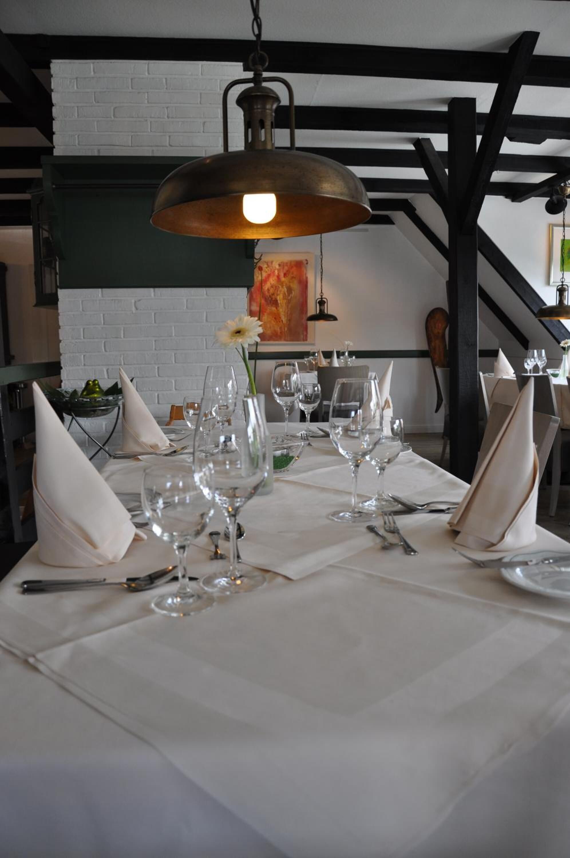A dining table set with white napkins, wine glasses, and cutlery, under a hanging light fixture.