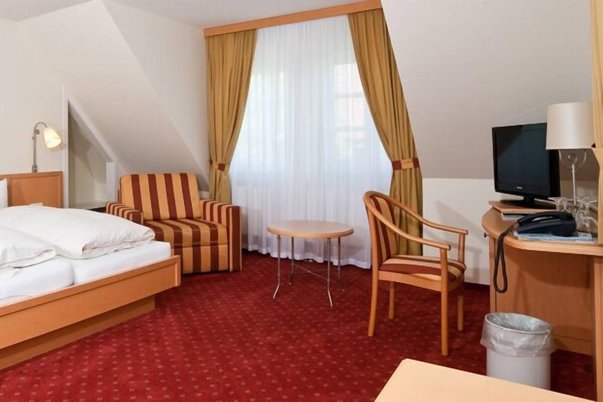 Ein Hotelzimmer mit einem Bett, einem Schreibtisch mit Computer, einem Sessel, einem kleinen Tisch und einer Mülltonne.