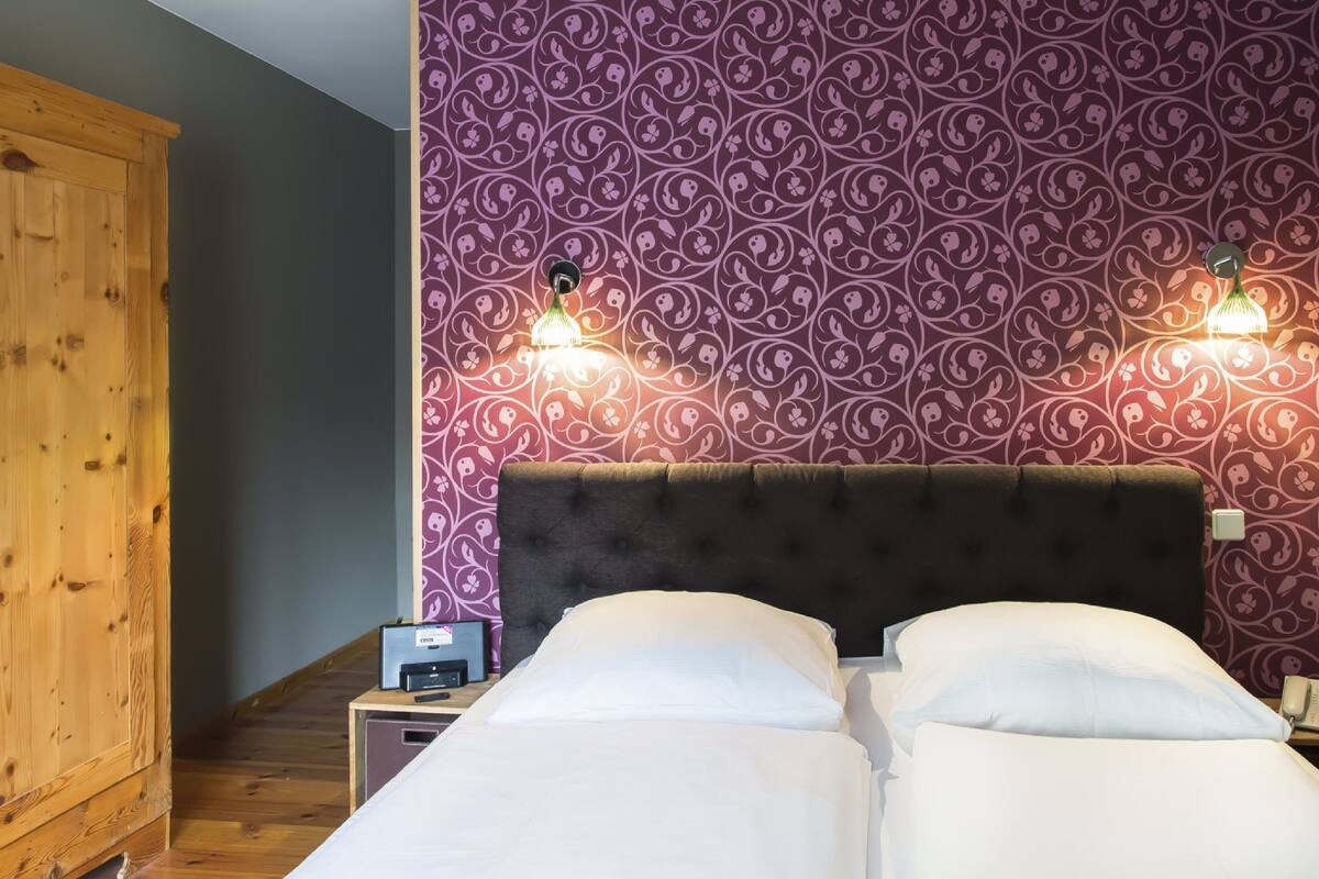 A bedroom with a wooden wardrobe, a bed with white bedding, and a patterned wall.