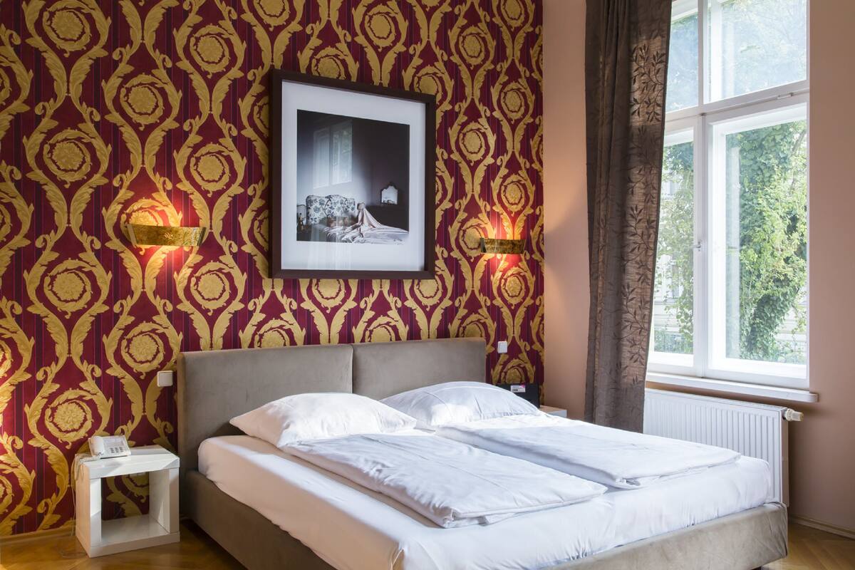 A neatly made bed in a room with patterned wallpaper, a framed picture on the wall, and a window with a view of greenery.
