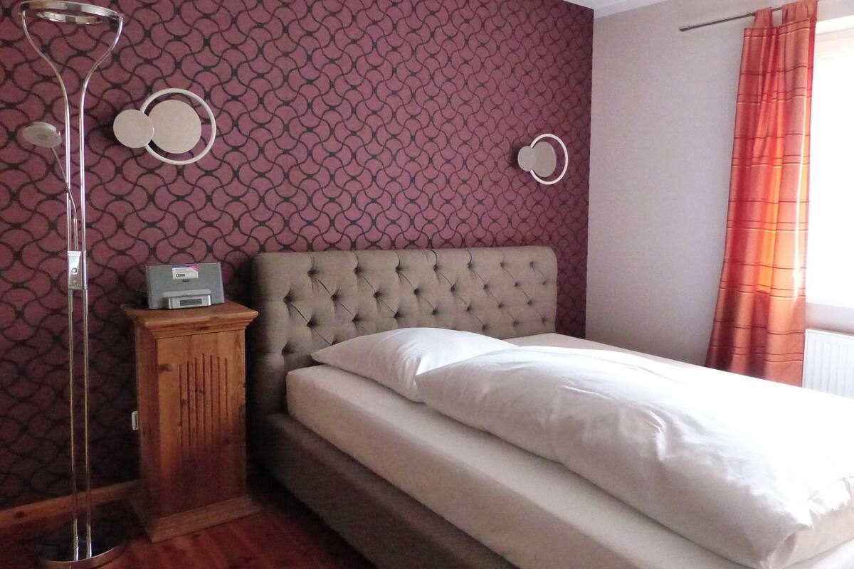 A bedroom with a tufted headboard, a bed with white linens, a wooden nightstand, a floor lamp, and a patterned wallpaper.