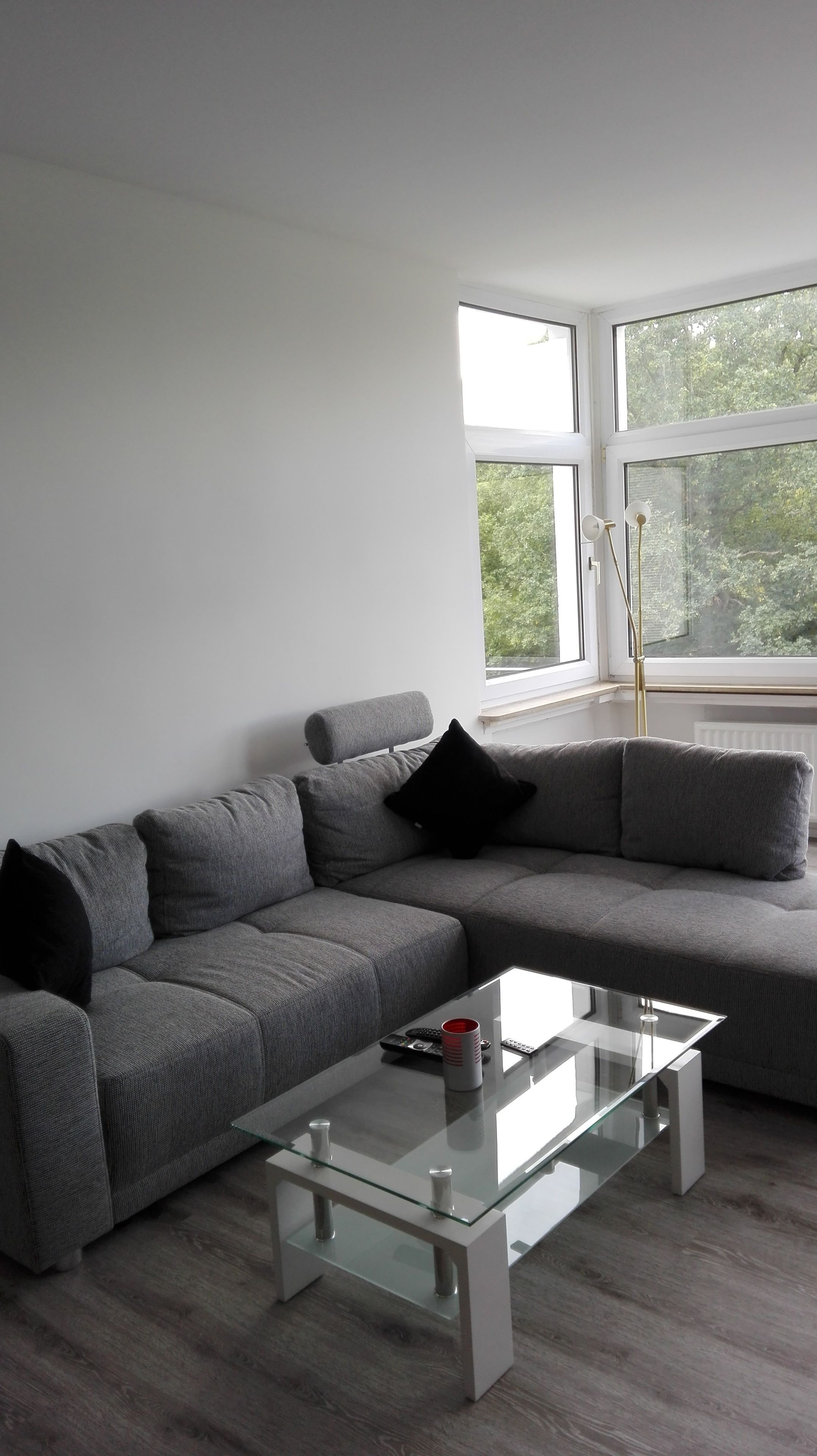 A modern living room with a grey sectional sofa, a glass coffee table, and a large window with a view of greenery.
