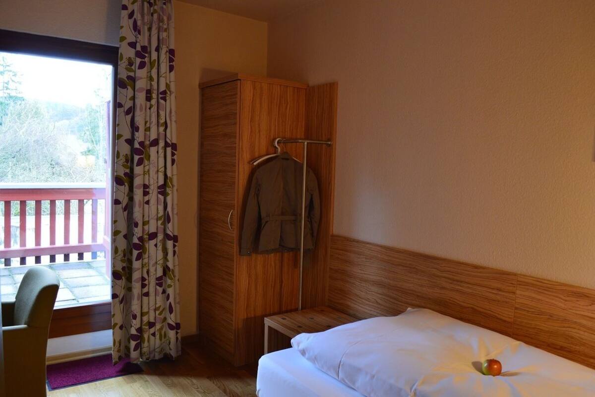 Hotellihuone, jossa on sänky, puinen vaatekaappi, tuoli ja ikkuna, josta avautuu näkymä puita kohti.