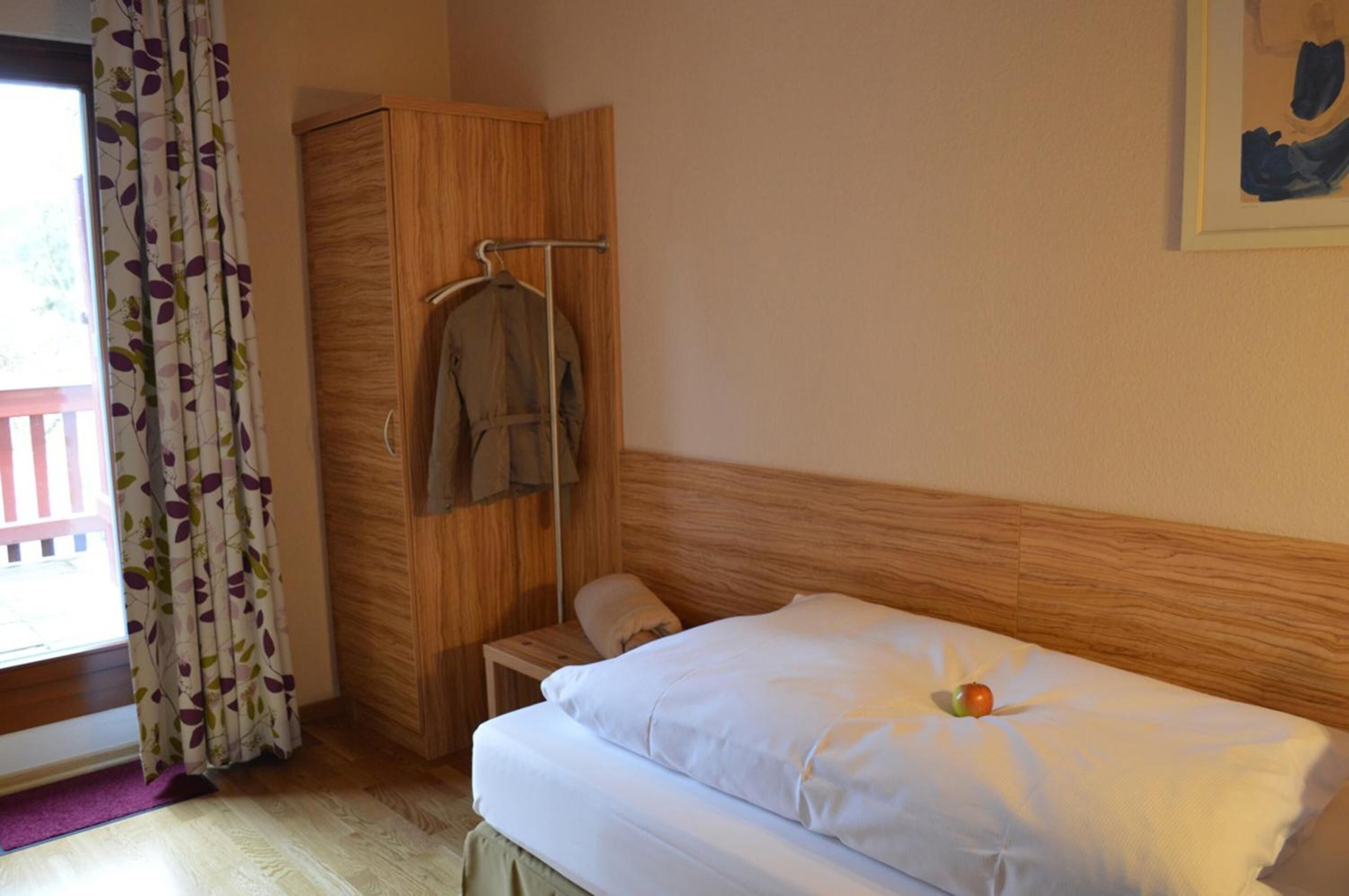 A bedroom with a wooden headboard, a bed with white bedding, a nightstand, a wardrobe, and a hanging coat rack.