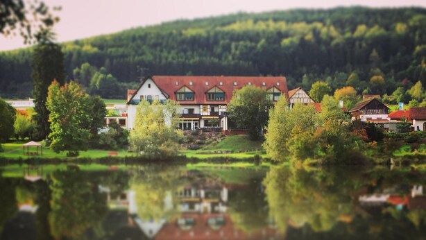 Una casa junto al lago con su reflejo, rodeada de árboles y una colina al fondo.