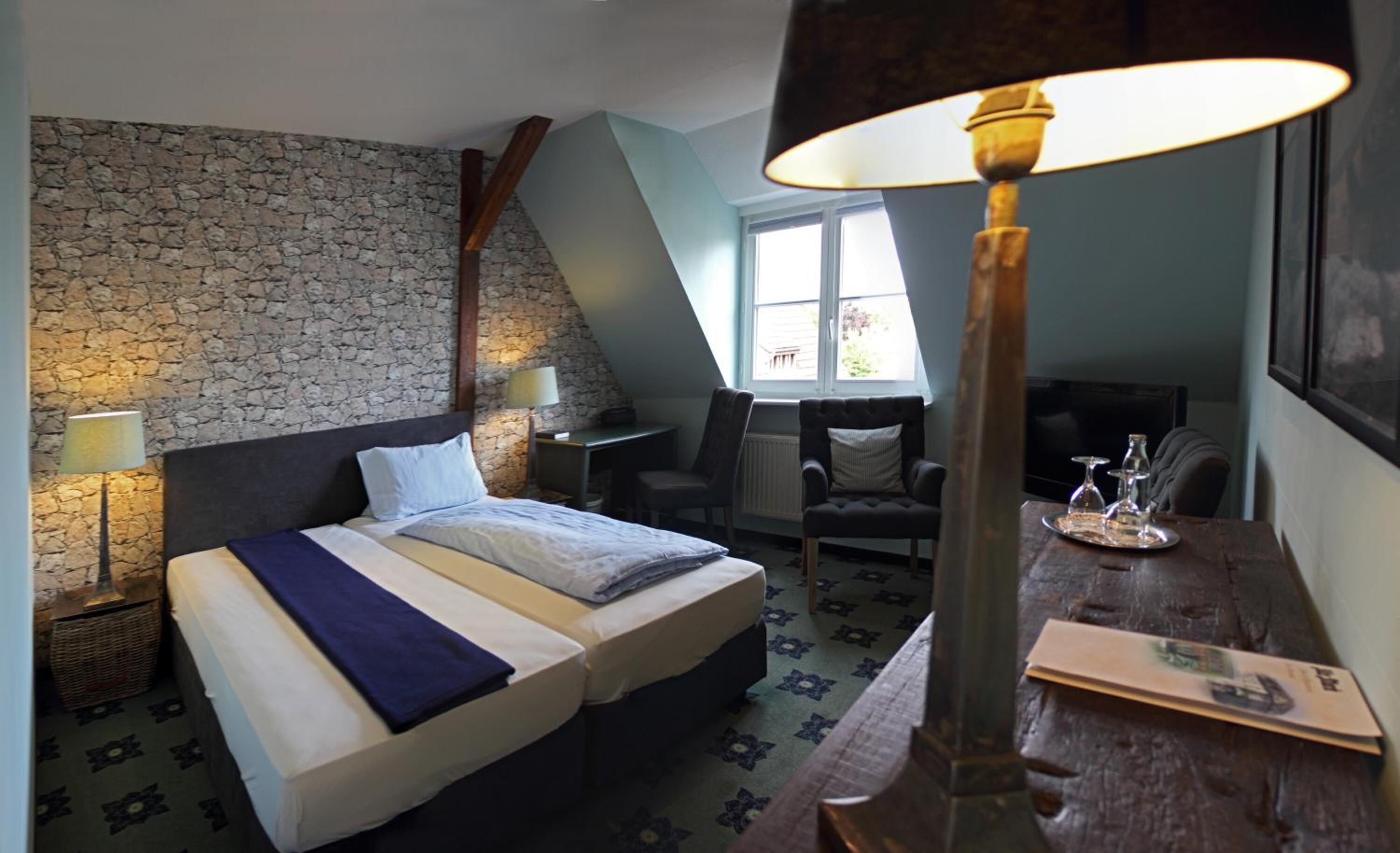 Une chambre avec un mur en pierre, un lit recouvert d’une couverture bleue, un bureau avec une lampe et une fenêtre donnant sur un beau paysage.