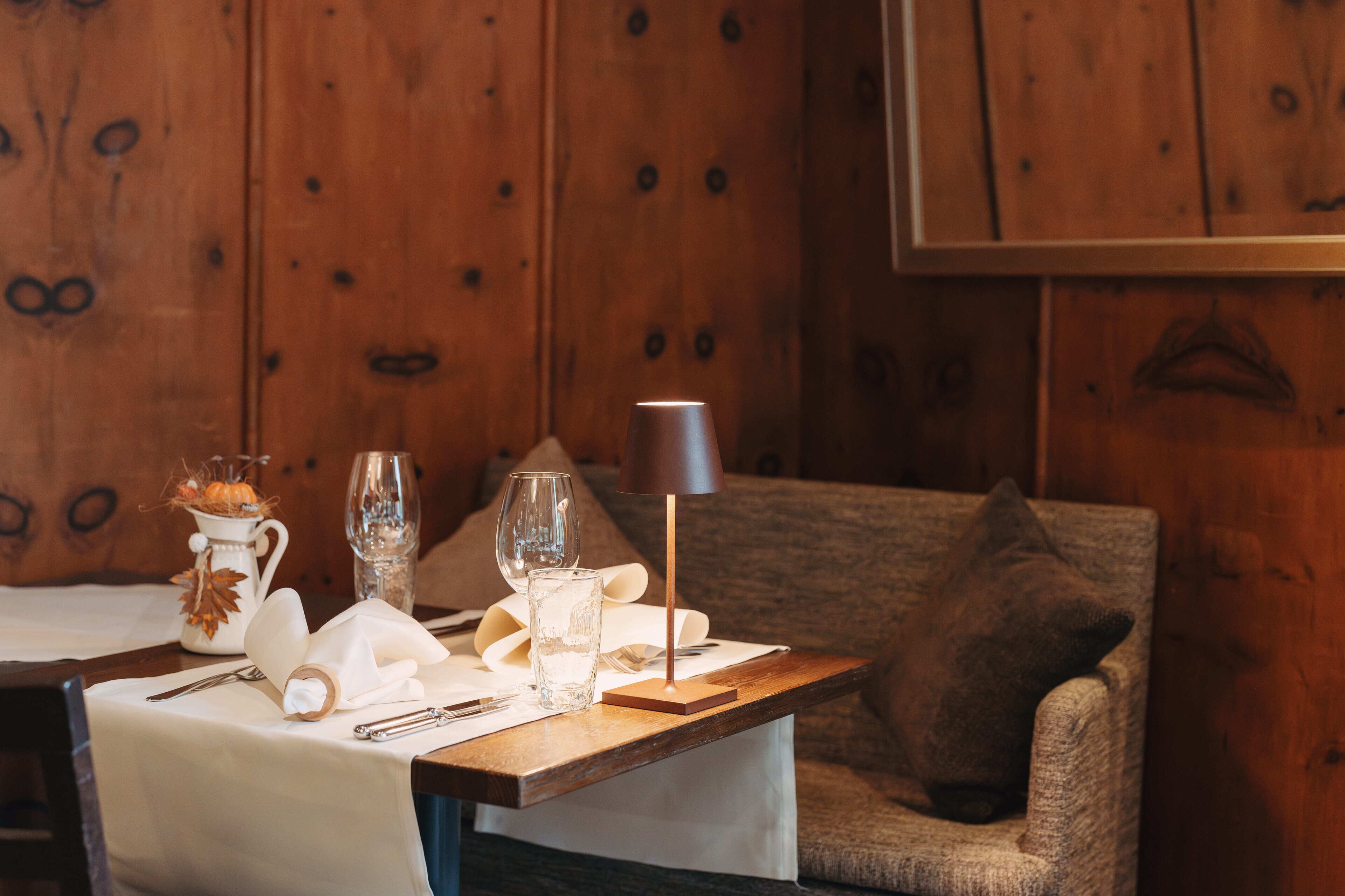 Une table à manger dressée avec une carafe, des verres et des couverts, une lampe et une chaise rembourrée, le tout dans une pièce en bois.