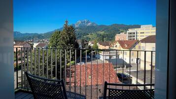 A balcony with metal railings, two black chairs, and a view of a town with buildings and mountains in the background.