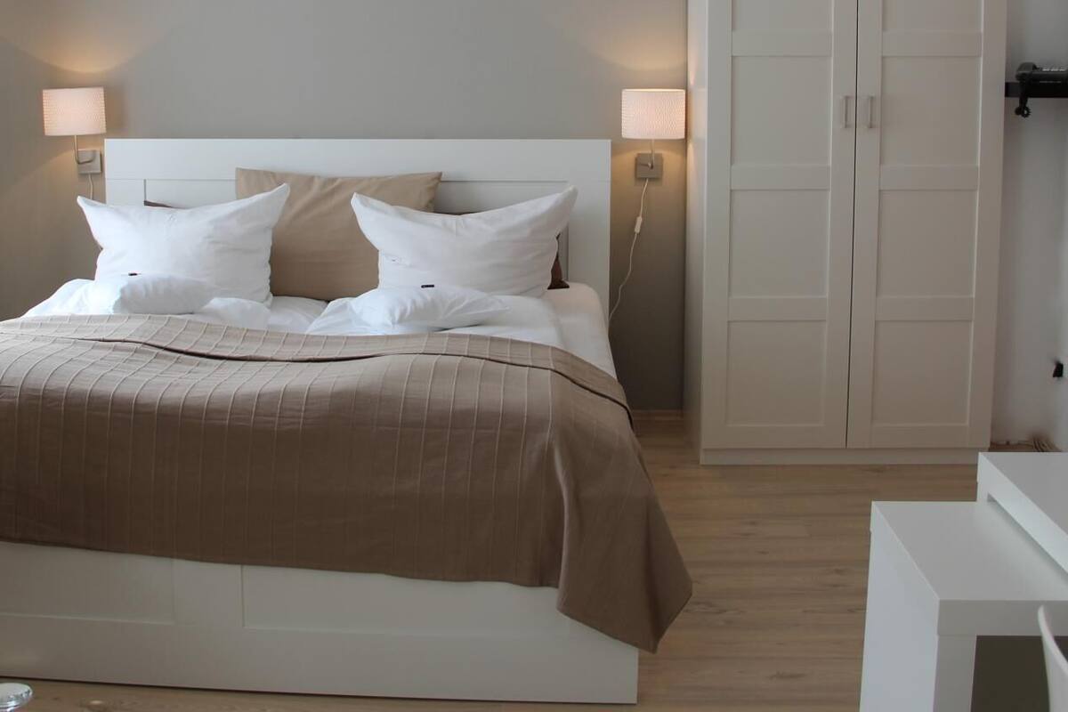 Een keurig opgemaakt bed met een beige sprei en witte kussens, een nachtkastje met een lamp en een inloopkast met schuifdeuren.
