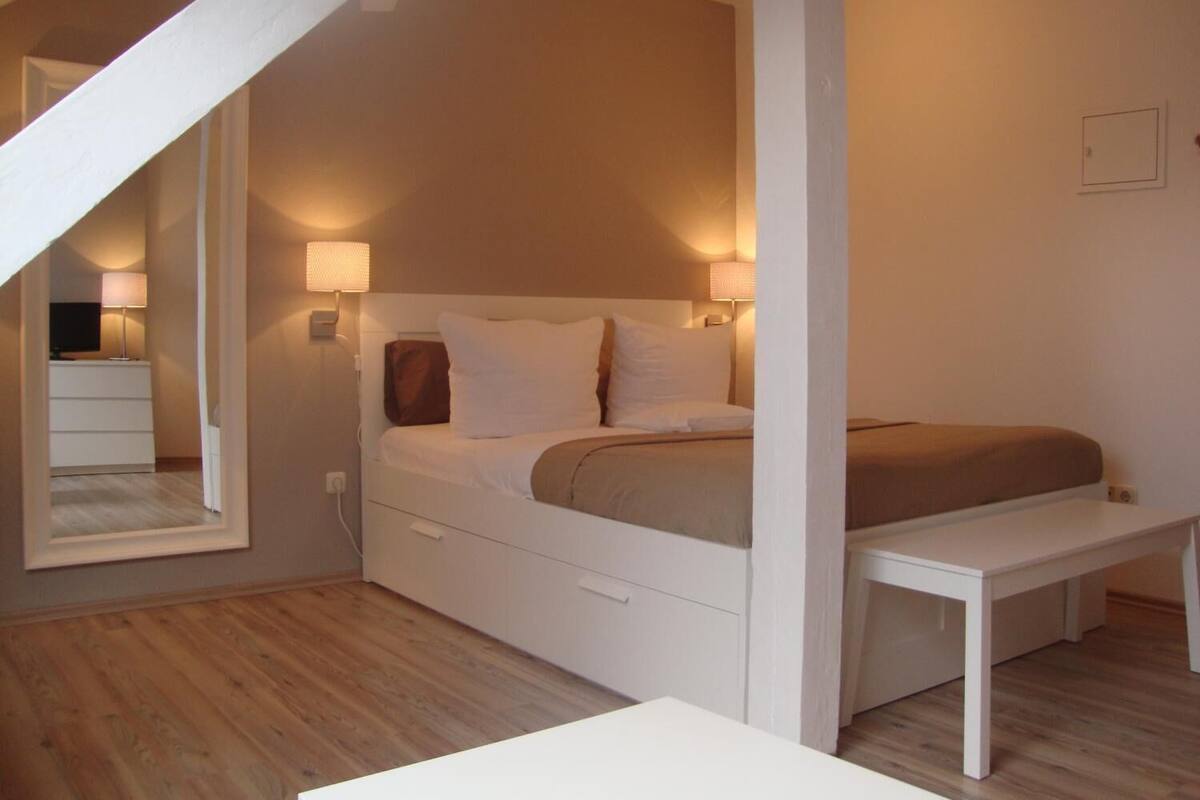 Een slaapkamer met een bed, nachtlampjes, een houten vloer, een witte bank en een spiegel.