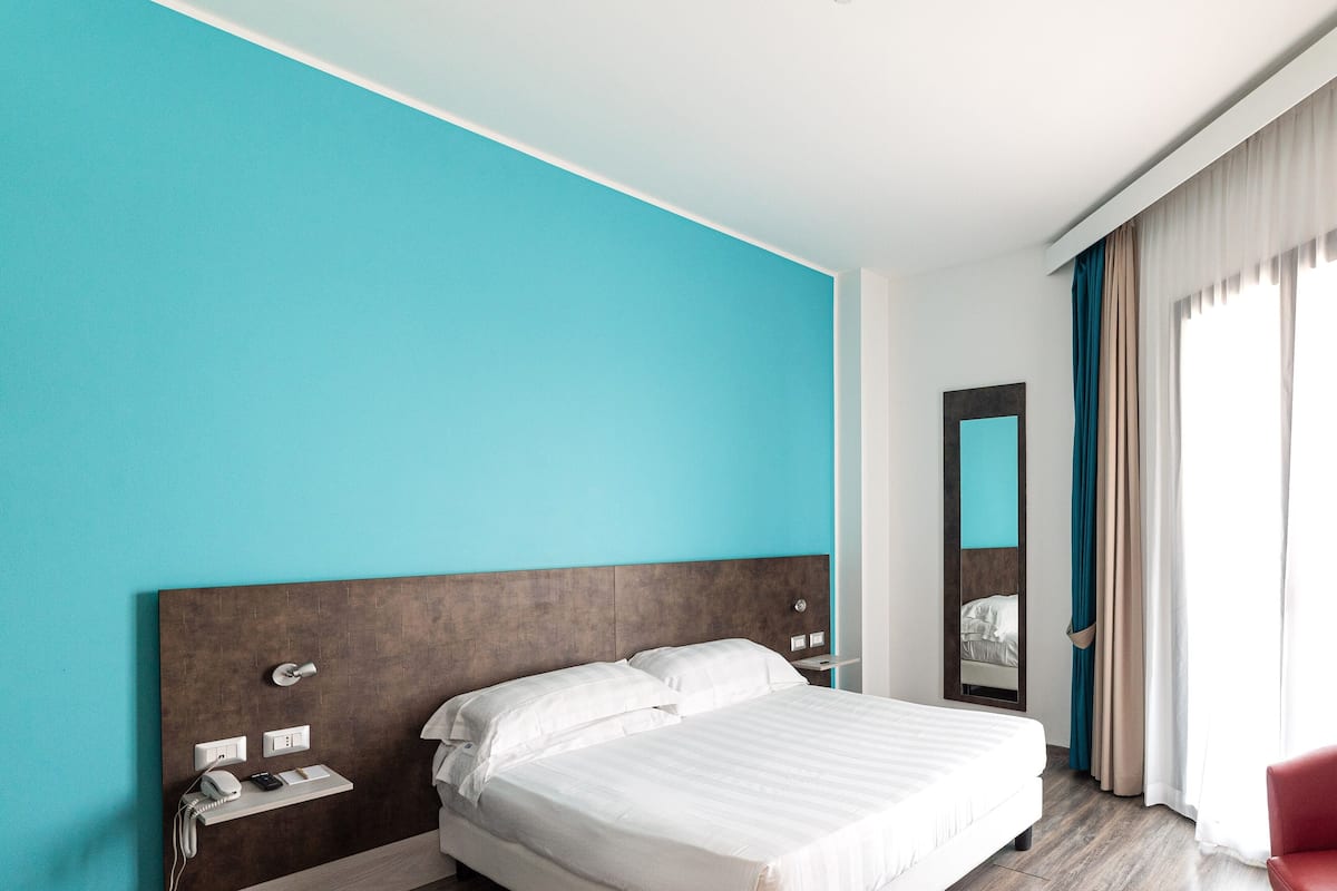Une chambre d’hôtel moderne avec un grand lit, une table de chevet, un miroir et un fauteuil rouge.