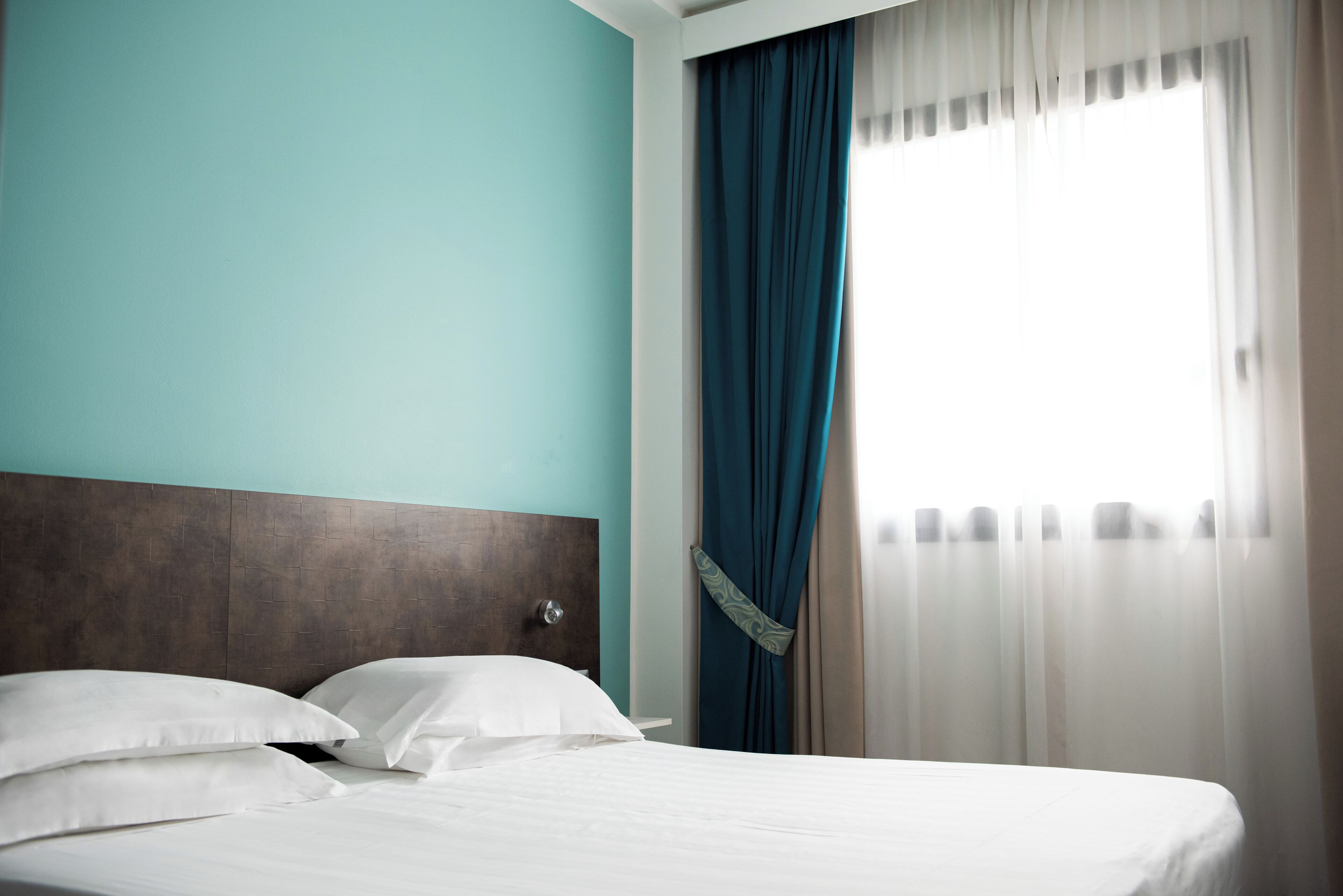 A hotel room with a bed, white pillows, a wooden headboard, teal walls, and sheer curtains.