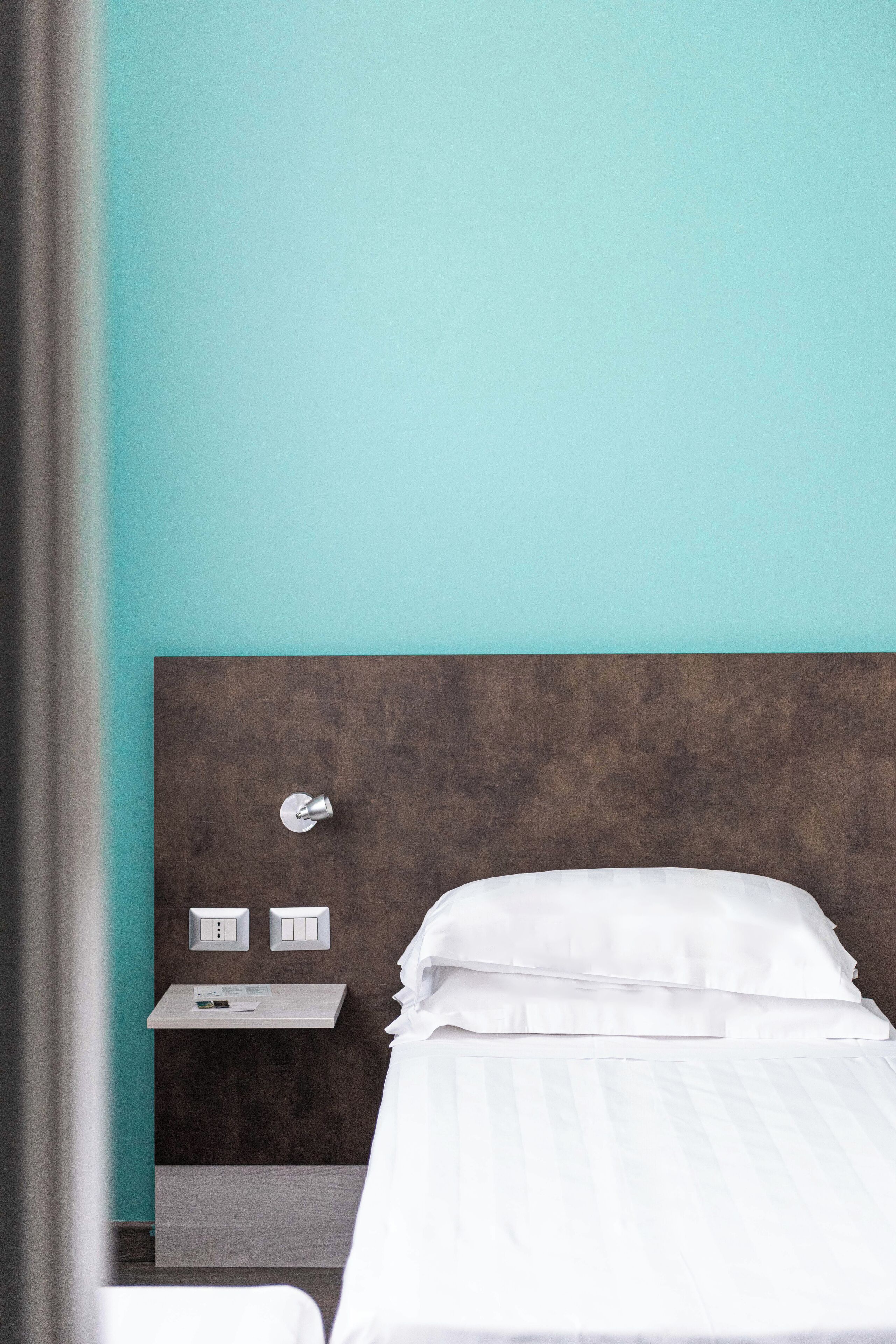 Une chambre d’hôtel avec un lit, une table de chevet, un interrupteur fixé au mur et une petite étagère sur laquelle se trouvent un livre et une télécommande.
