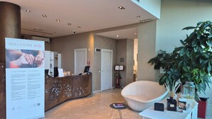 A reception area with a 'Welcome Aid' sign, a reception desk, a white chair, a table with amenities, and a large potted plant. - Phi Hotel Milano (Baranzate)