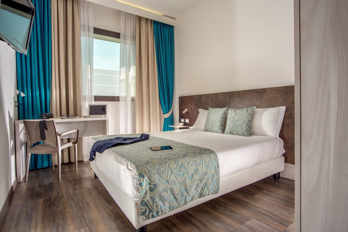 A hotel room with a bed, a desk, a chair, a TV, and a window with curtains.