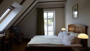 Een slaapkamer met schuine wand, een bed met witte lakens, een bureau met stoel en een raam met gordijnen.