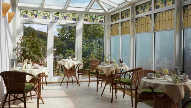 A conservatory with glass walls, wicker furniture, and a view of greenery outside. - Hotel Snorrenburg (Burbach)