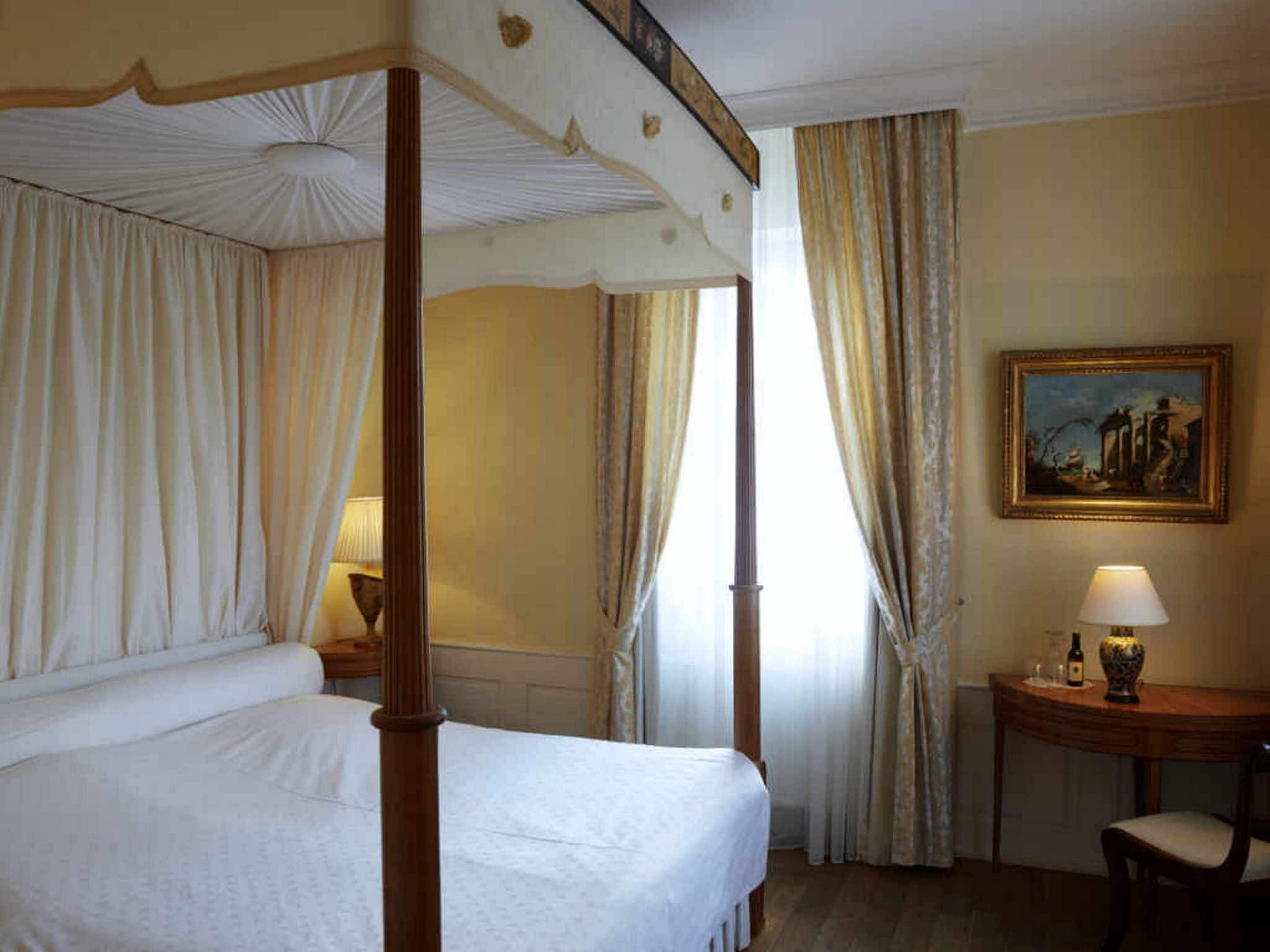 Ein Bett mit Baldachin, ausgestattet mit weißen Leinen, ein Nachttisch aus Holz mit Lampe, ein kleiner Tisch ebenfalls mit Lampe und ein Gemälde an der Wand.