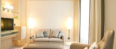 A modern hotel room with a sofa, armchair, and a flat-screen TV on a glass shelf.