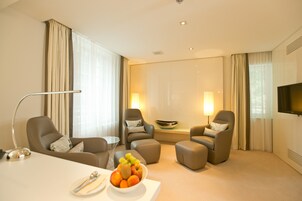 Una habitación de hotel moderna con una cama grande, dos sillones, una pequeña mesa con fruta y un televisor en la pared.