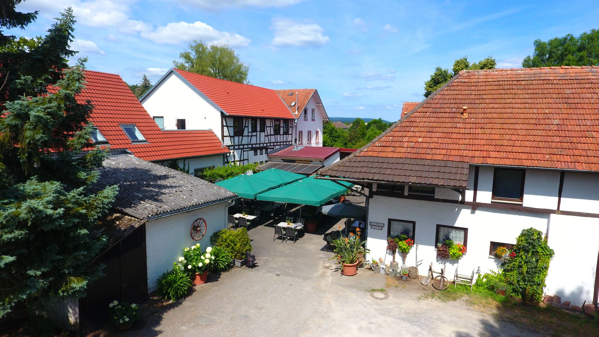 Ein Innenhof mit traditioneller europäischer Architektur, darunter ein Fachwerkhäuschen und ein Gebäude mit rotem Ziegeldach. Unter einem grünen Markisenstoff befinden sich Tische und Stühle im Freien, was auf einen Gastbereich hindeutet.