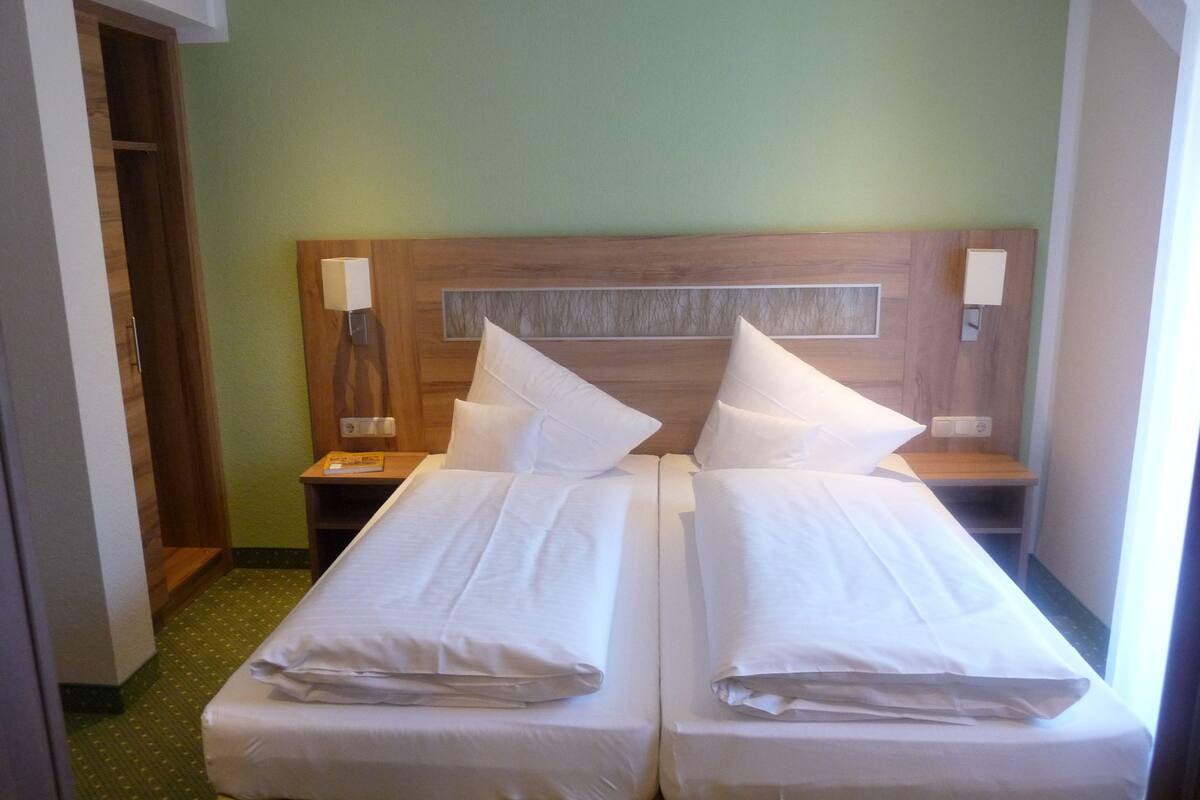 A hotel room with two single beds, wooden headboards, bedside tables, and a green carpet.