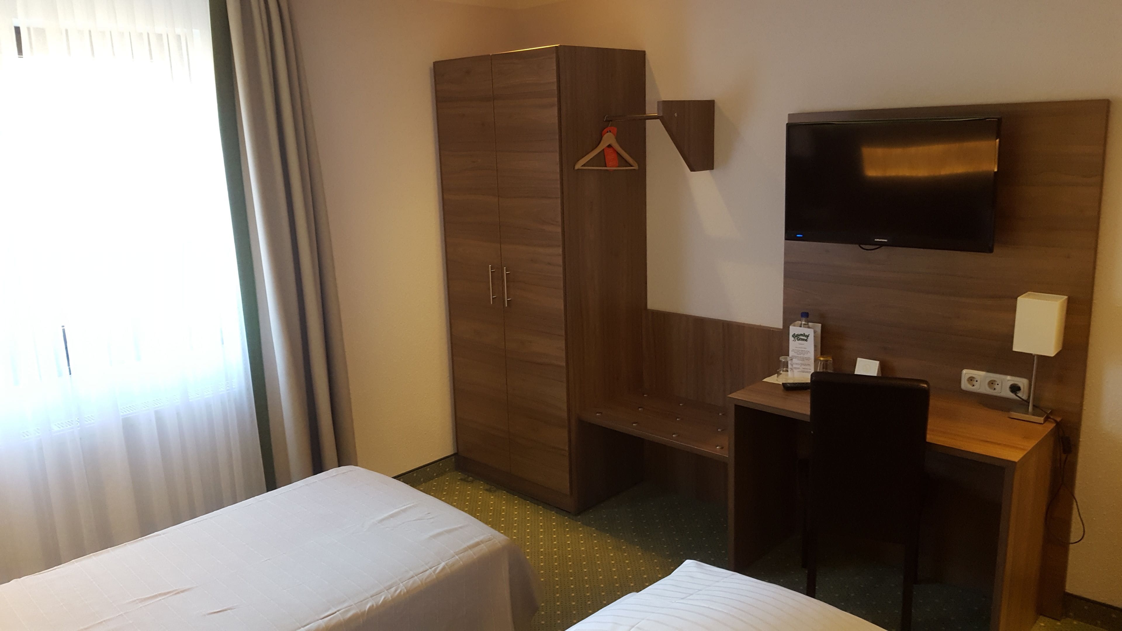 A hotel room with a bed, a desk with a chair, a wardrobe, and a TV mounted on the wall.