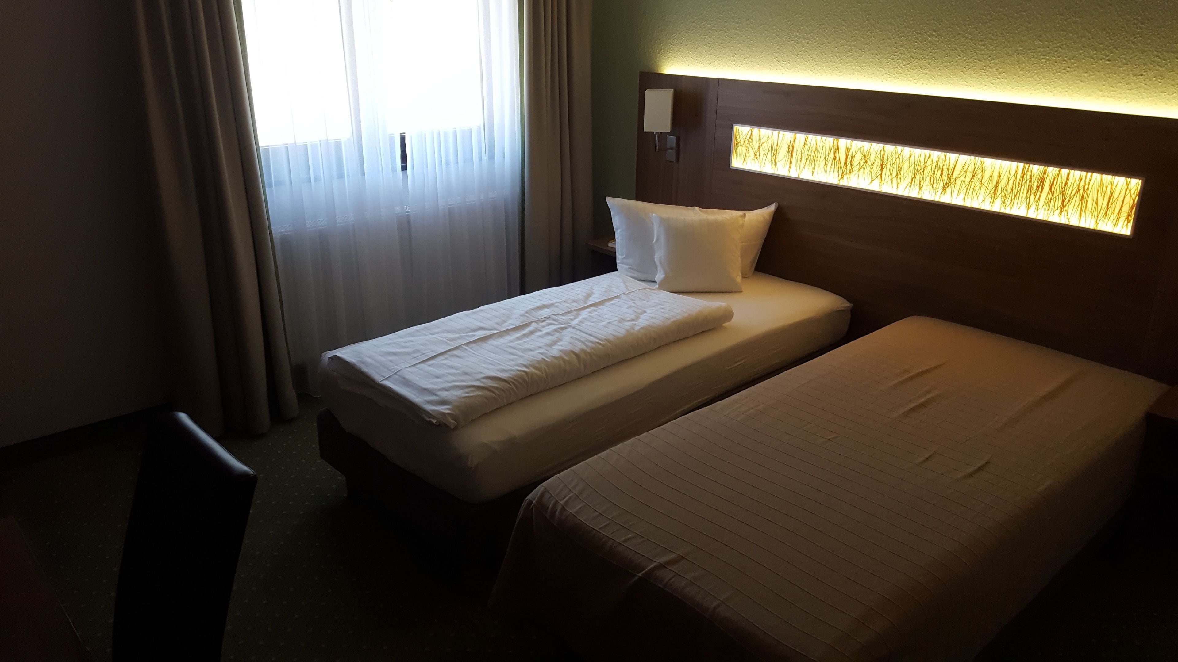 Ein Hotelzimmer mit zwei Einzelbetten, einem Nachttisch, einer Lampe und einem Fenster mit Vorhängen.
