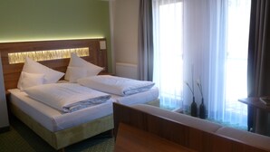 Una cama doble con sábanas blancas, un cabecero de madera y una mesita de noche. Se ve una ventana con cortinas traslúcidas y vistas al exterior.