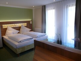 Una cama doble con sábanas blancas, un cabecero de madera y una mesita de noche. Se ve una ventana con cortinas traslúcidas y vistas al exterior.