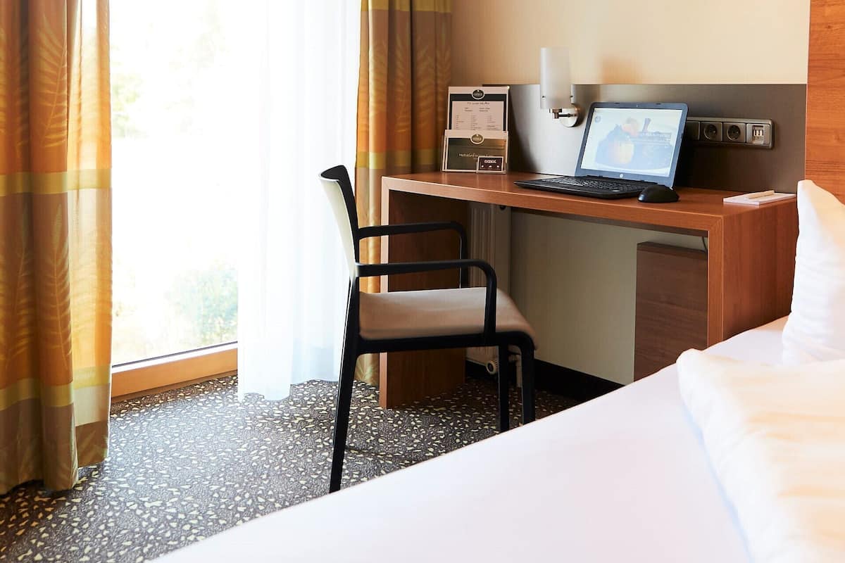 A hotel room with a bed, a desk with a laptop, a chair, and a window with curtains.