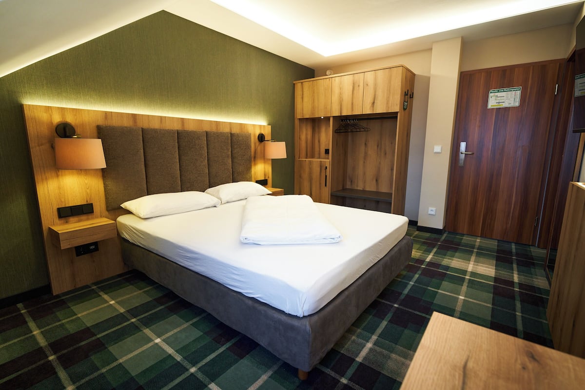 A hotel room with a bed, a wooden wardrobe, a nightstand with a lamp, and a plaid carpet.