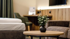 Et hotelværelse med seng, sofa, lille rundt bord med blomster, et fjernsyn monteret på væggen og en receptionsdisk i baggrunden.