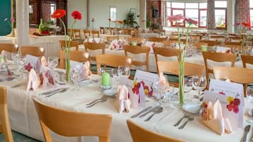Uma sala de banquetes com várias mesas montadas para um evento formal, cada uma com louça, copos e arranjos florais.