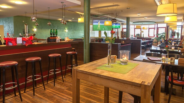 A restaurant interior with wooden tables, bar stools, hanging pendant lights, and a television screen in the background. - Sporthotel Öhringen (Öhringen)