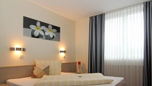 A hotel room with a bed, two bedside tables, a nightstand, a painting, and a window with curtains. - Sporthotel Öhringen (Öhringen)