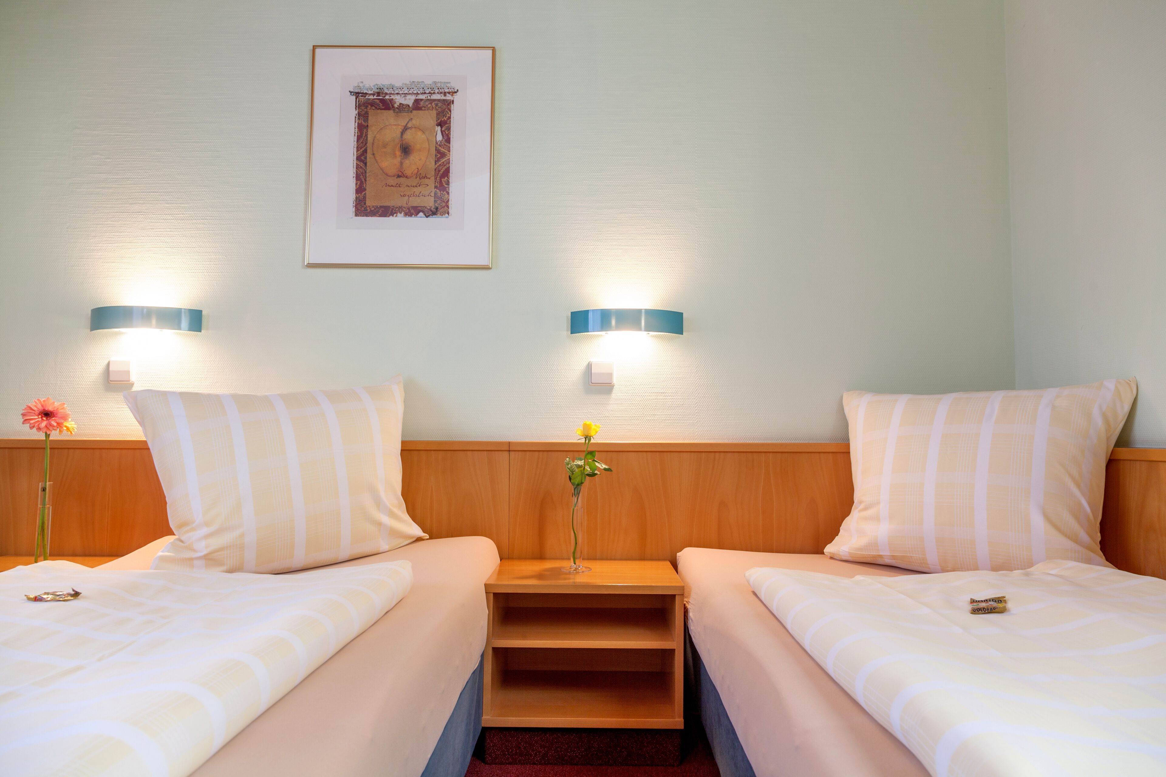 Una habitación de hotel con dos camas, cada una con un cojín y una mesita de noche. Hay un cuadro enmarcado en la pared sobre las camas.