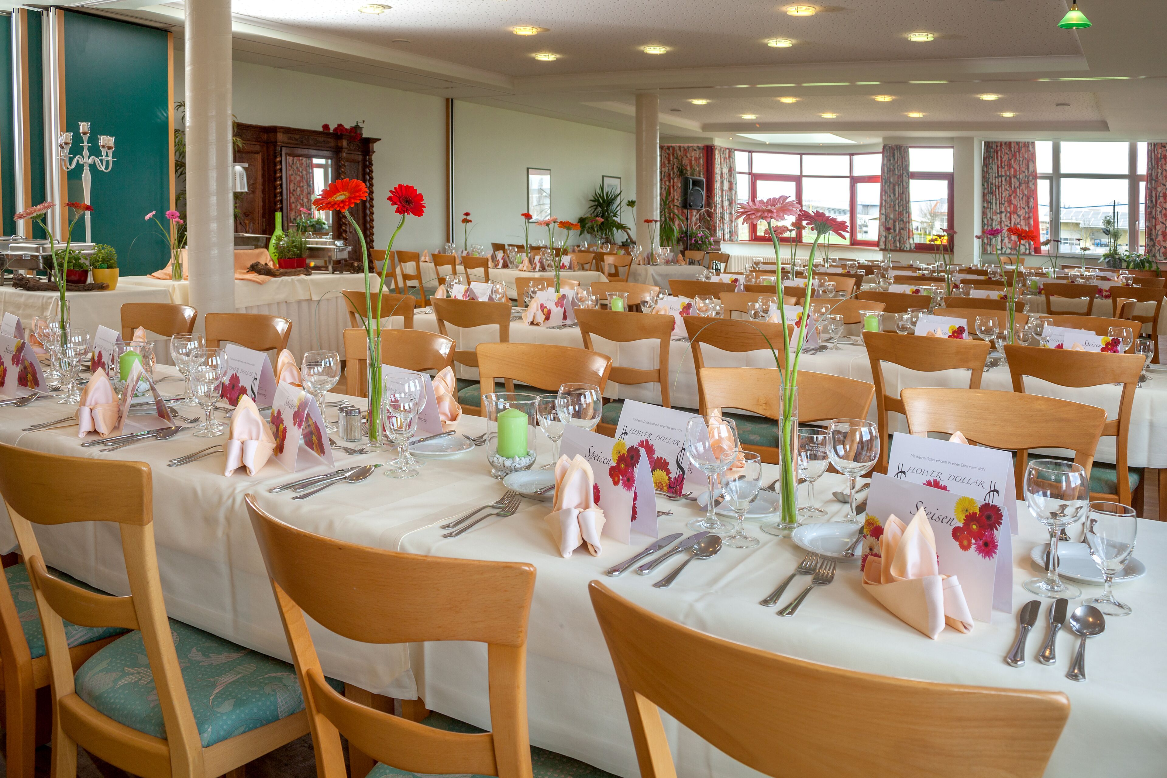 A large dining hall with multiple tables set for a formal event, featuring wooden chairs and white tablecloths.